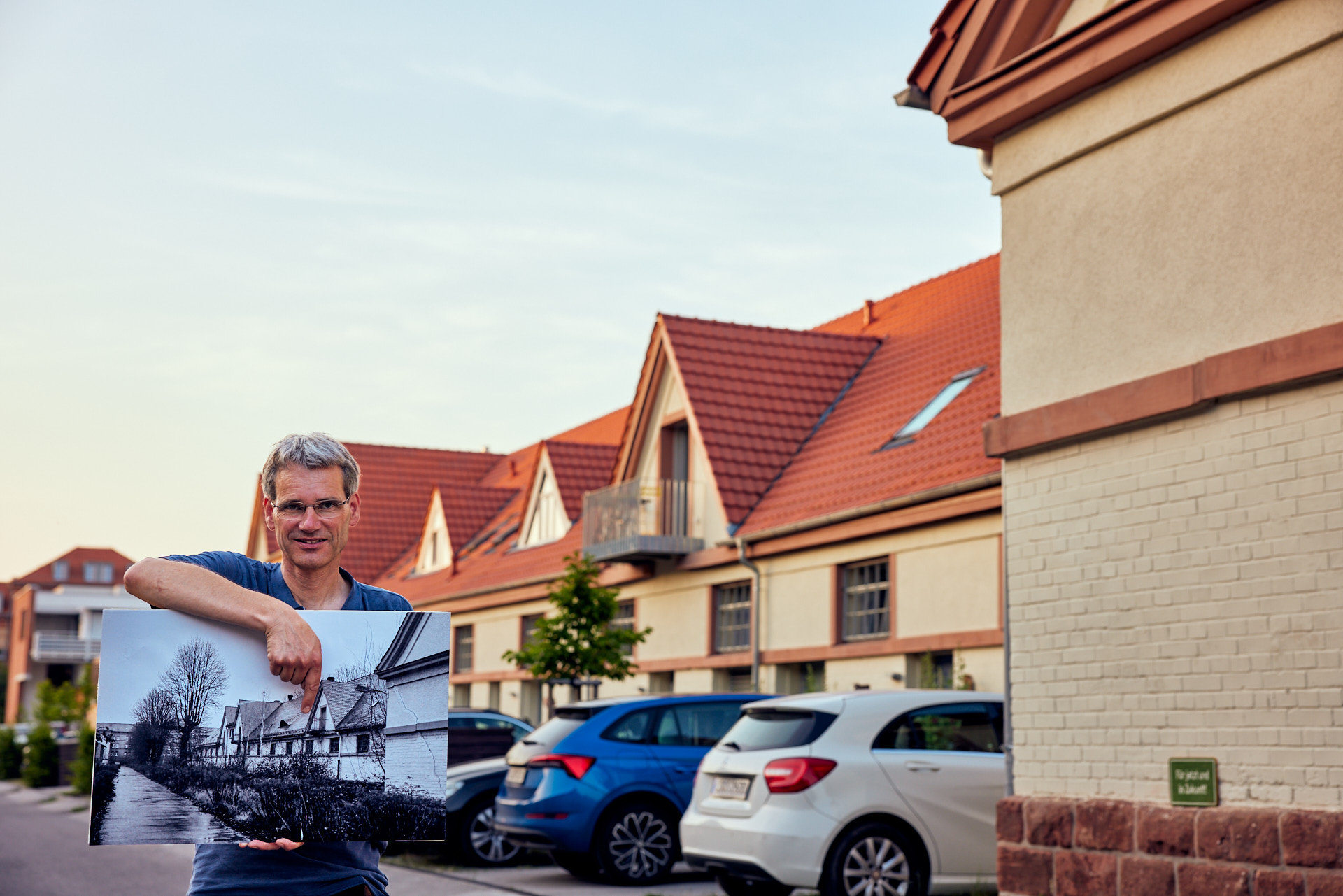 Peter Eichenlaub mit Vorherbild vor den zu Wohnungen umgebauten alten Militär-Pferdeställen mit Schild „Für jetzt und in Zukunft!“ (Foto: Niko Martin)