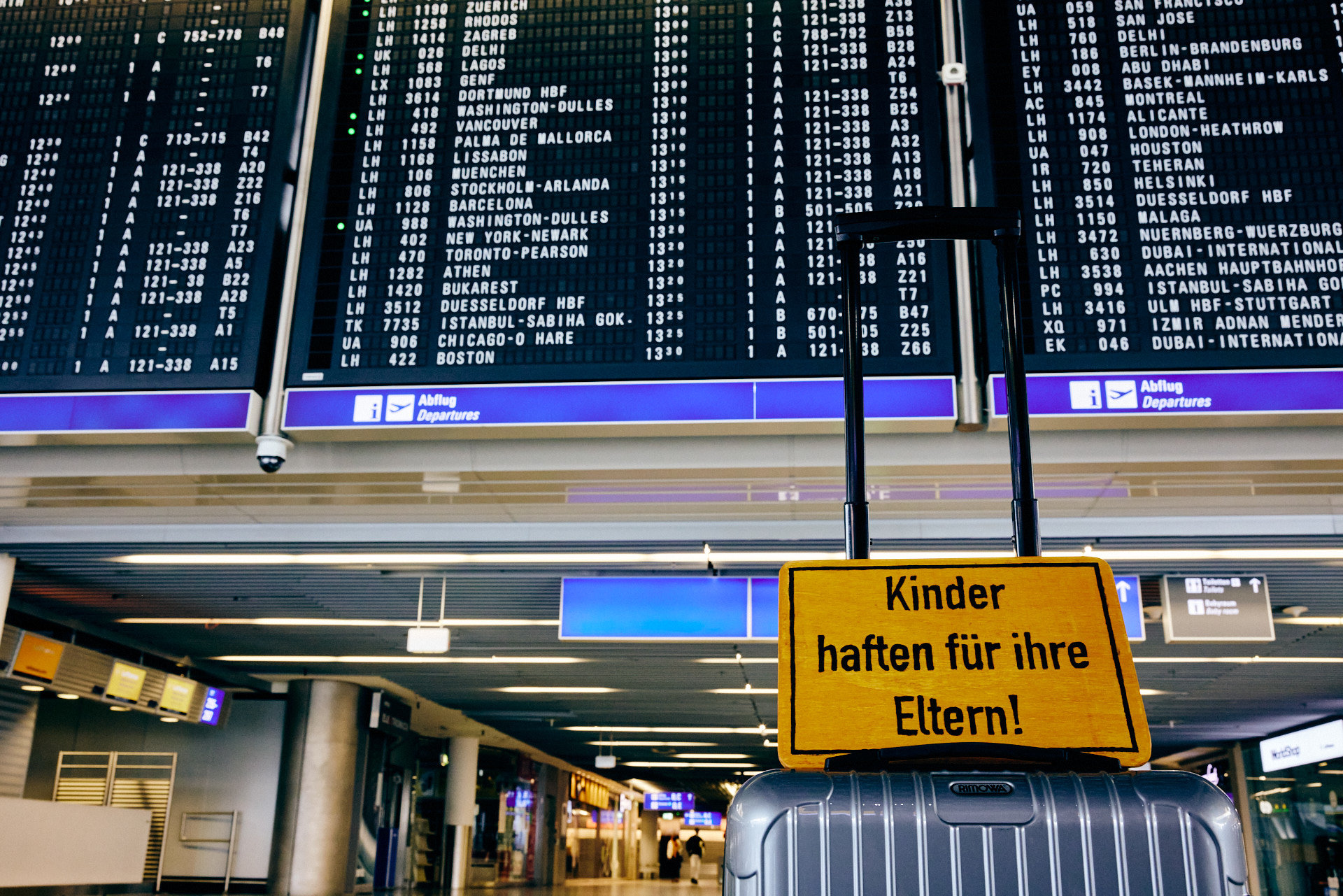 Abflugtafel FRA – Frankfurt Airport mit Schild „Kinder haften für ihre Eltern!“ (Foto: Niko Martin)